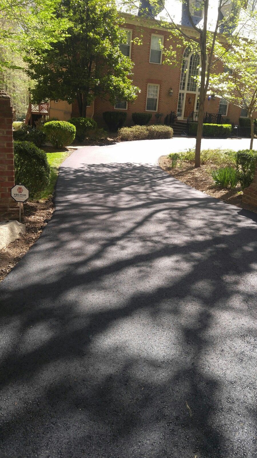 Freshly paved asphalt driveway leading to a two-story brick house with mature trees and landscaping.