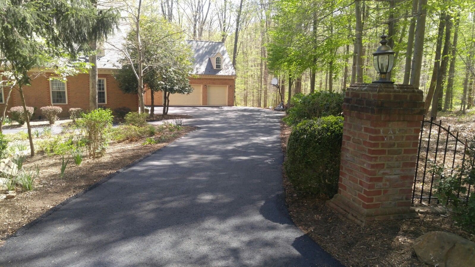 A paved driveway leads toward a brick house with an attached garage, framed by trees and a brick pillar with a lamp.
