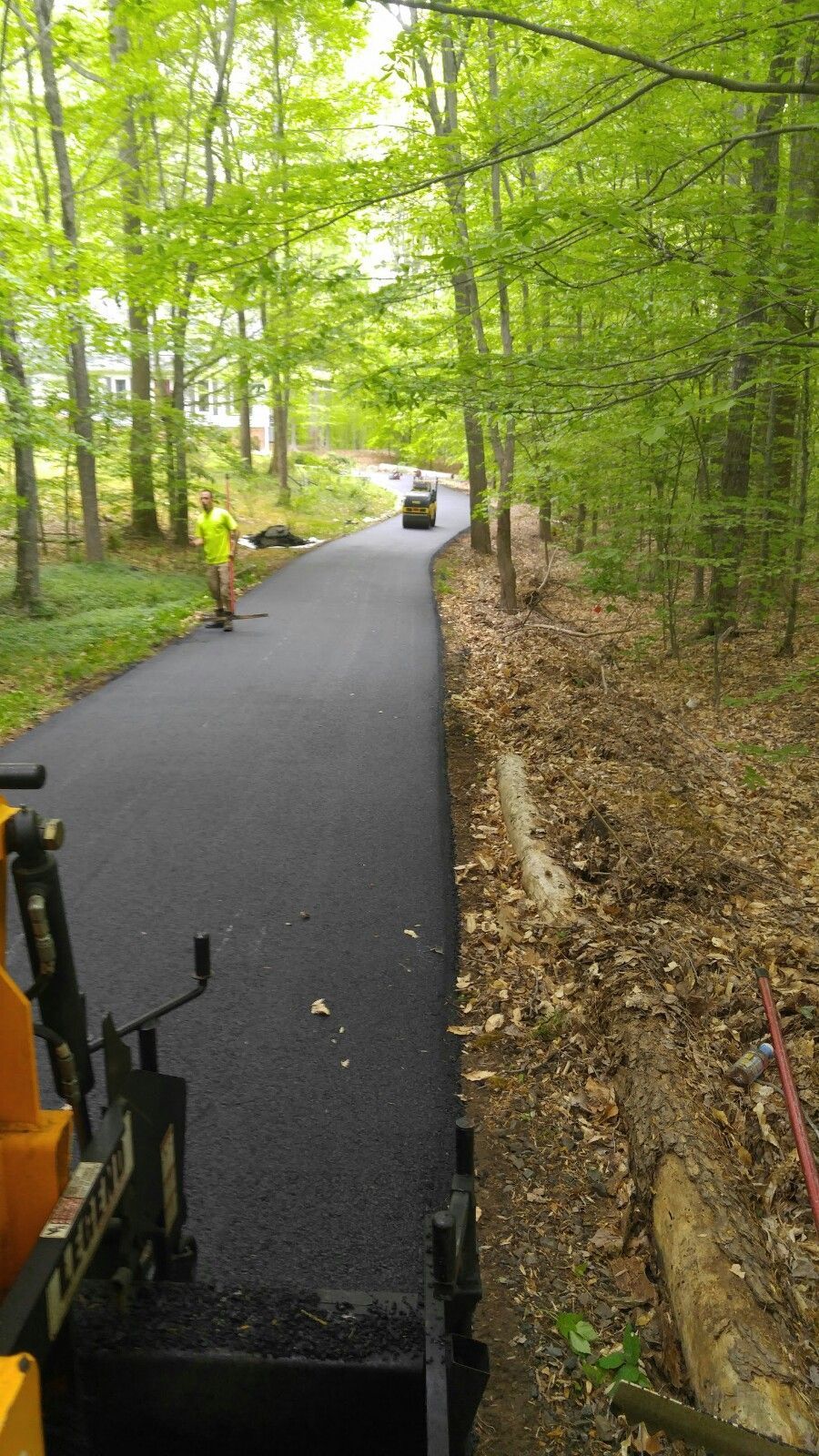 A worker in a yellow vest paving a long, fresh asphalt road winding through a lush green forest.