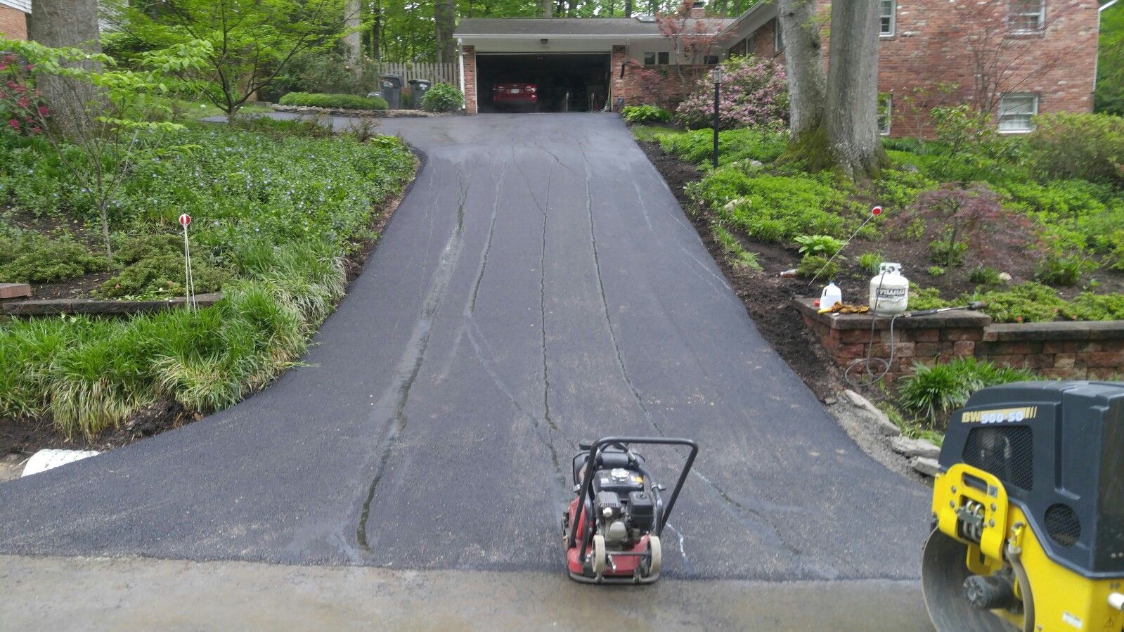 A freshly paved dark asphalt driveway leads to a residential garage, with a small motorized roller parked in the foreground.