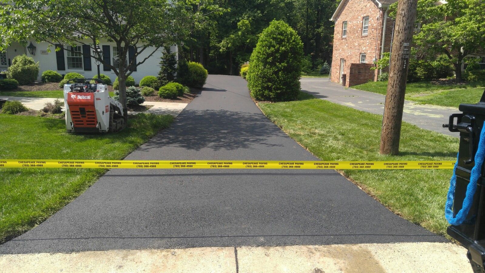 A freshly paved asphalt driveway cordoned off with yellow caution tape, with a small construction vehicle parked nearby.