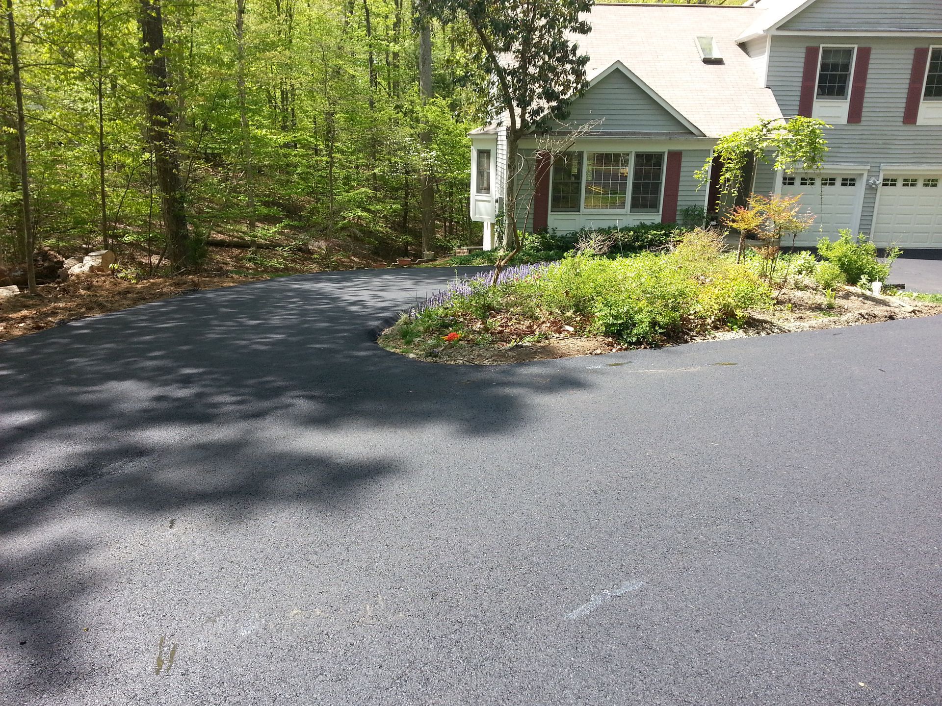 A freshly paved dark asphalt driveway curves in front of a house next to a wooded area and a small garden bed.