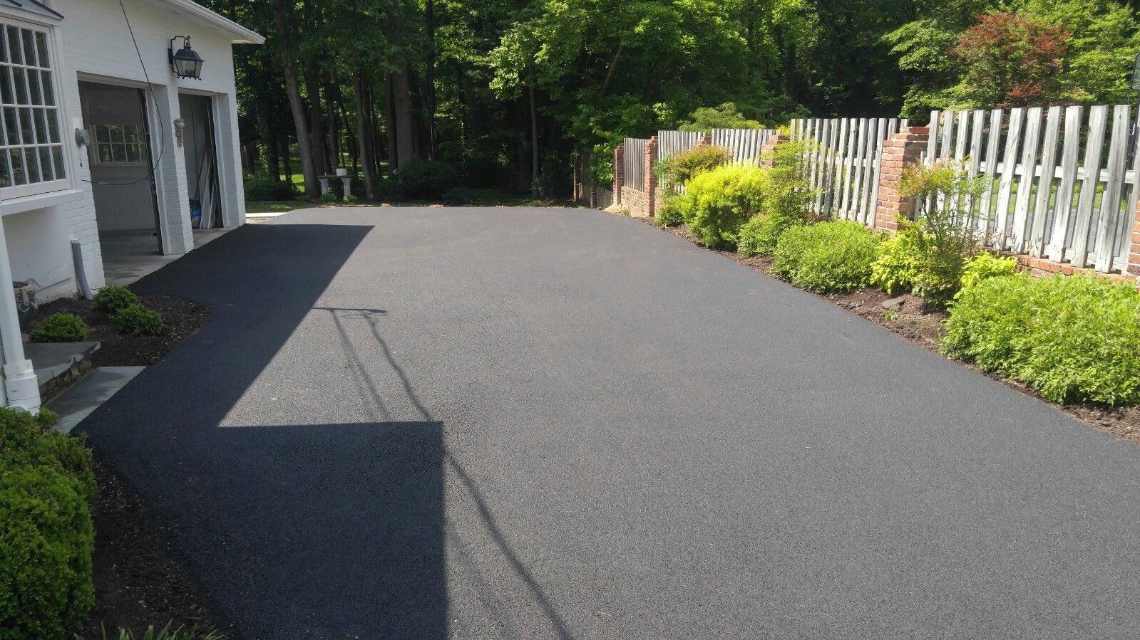 A newly paved dark asphalt driveway leads to a garage, bordered by a white picket fence and green landscaping.