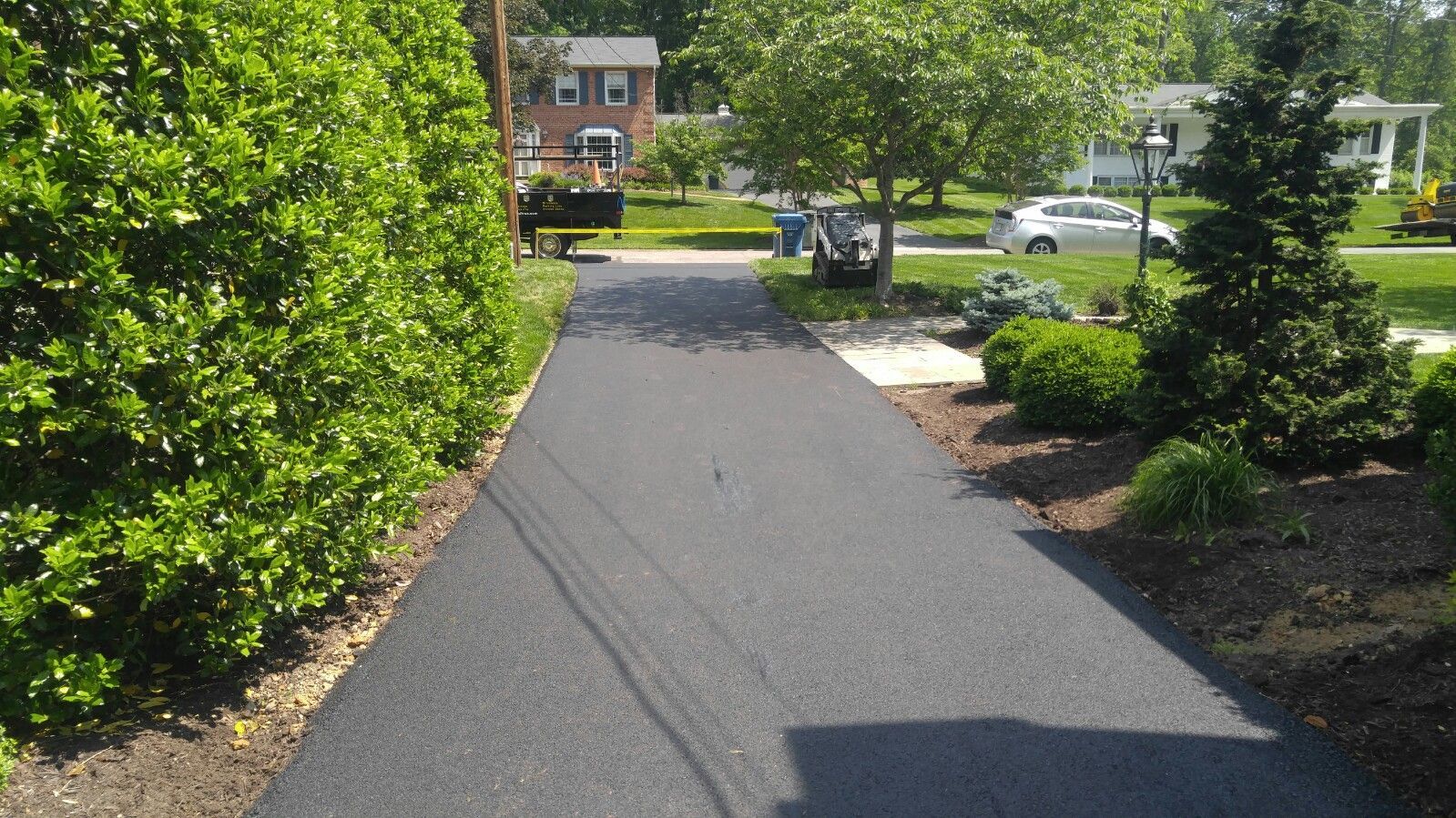 A newly paved black asphalt driveway stretches between a lush green hedge and a landscaped garden with a small evergreen.