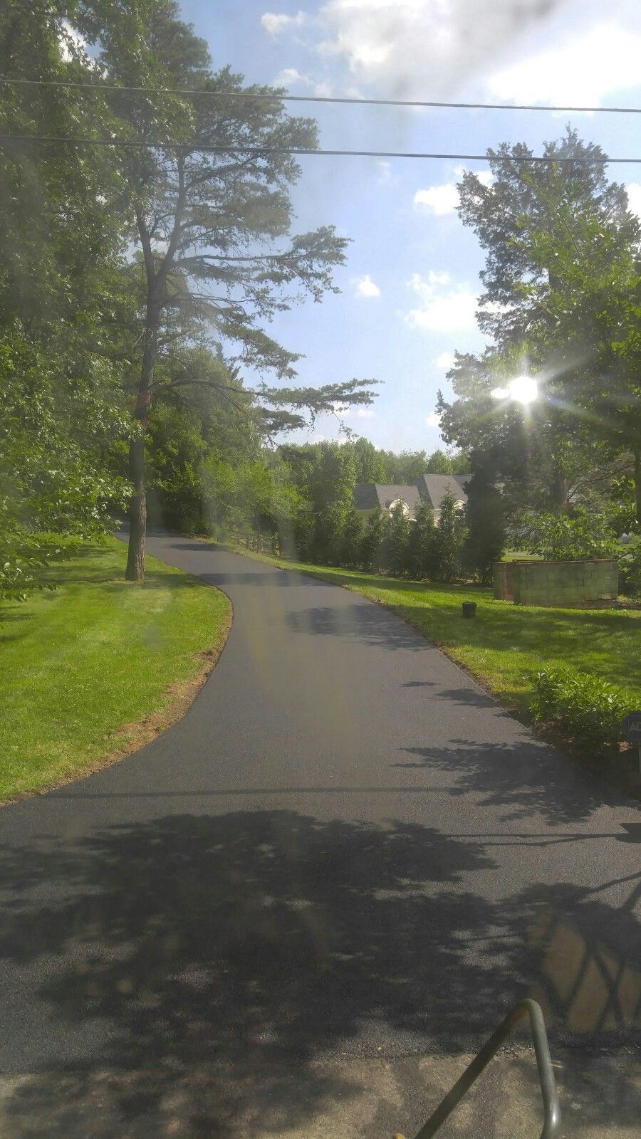 A paved driveway winds through a green, tree-lined yard under a bright, sunny sky with lens flare.