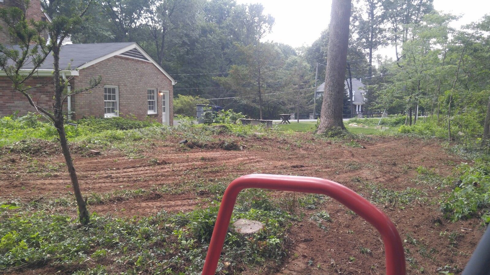 A red frame in the foreground overlooks a yard with cleared, exposed red soil and a brick house in the background.