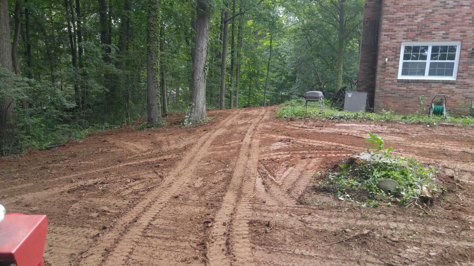 Muddy ground with heavy tire tracks leading toward trees and the side of a brick house.
