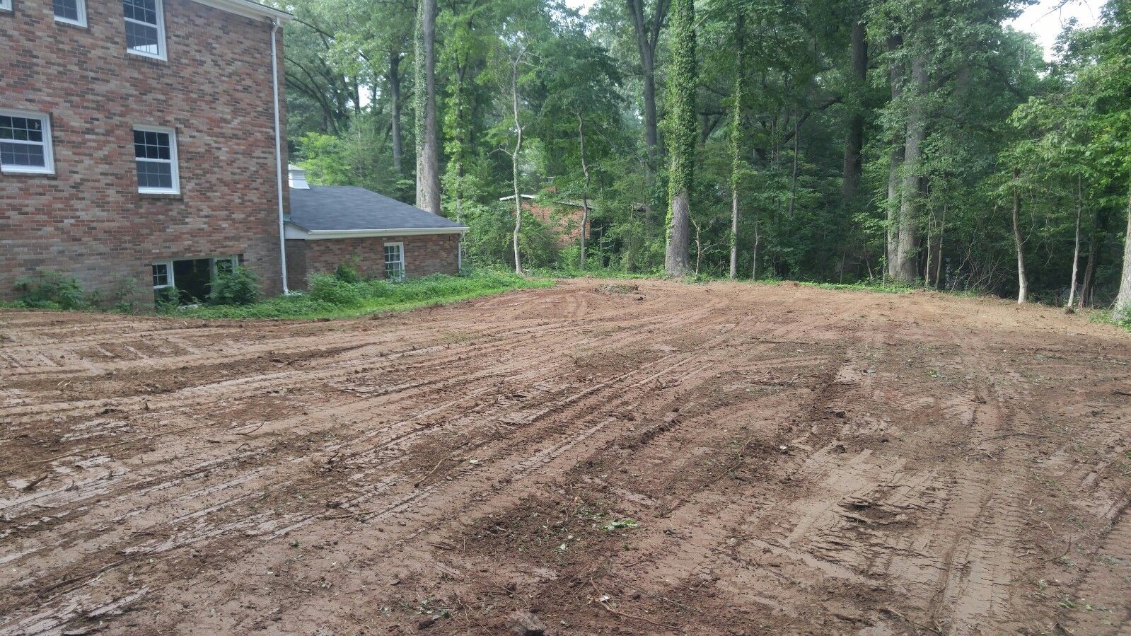 A brick house stands next to a large, cleared plot of dirt earth surrounded by dense trees.