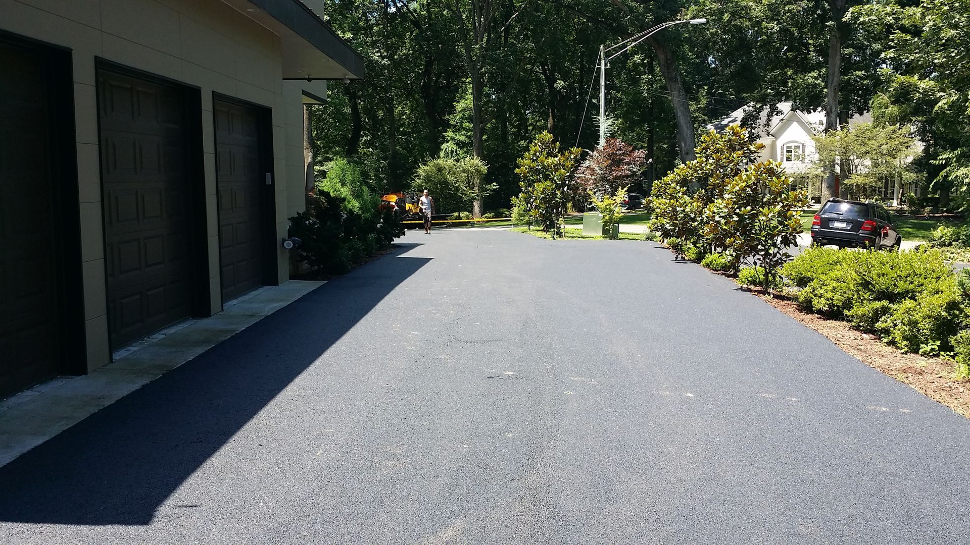 A newly paved, dark asphalt driveway leads to a three-car garage beside a house with green trees and landscaping.