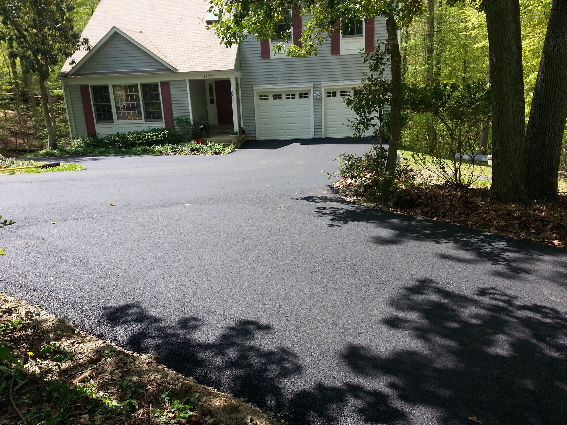 A freshly paved, dark asphalt driveway leading to the two-car garage of a suburban house surrounded by trees.