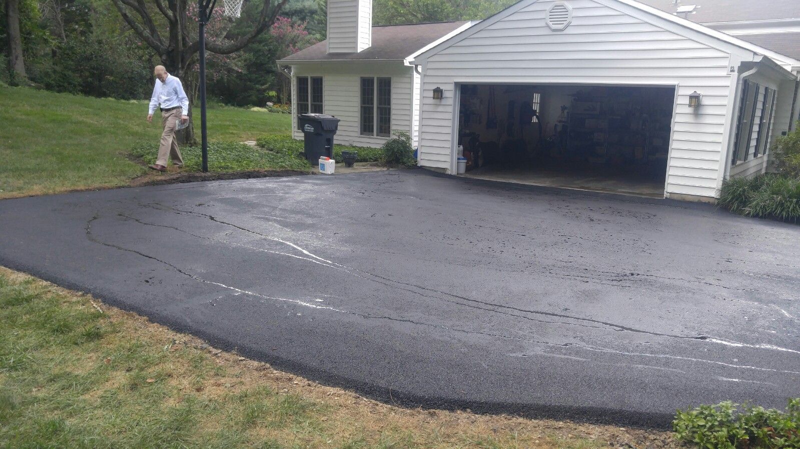 A man walks on the grass next to a freshly paved black asphalt driveway leading to a white suburban garage.