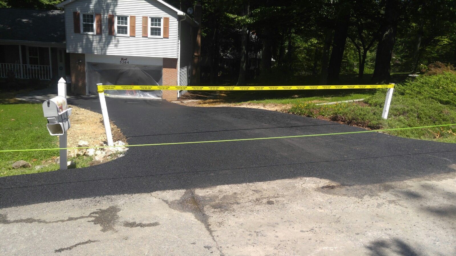 A freshly paved asphalt driveway blocked off with yellow caution tape in front of a gray house.