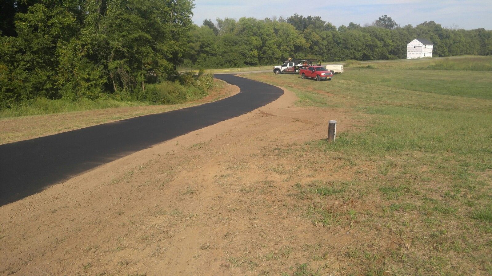A freshly paved black asphalt path curves through a grassy field toward trees, with vehicles parked in the distance.