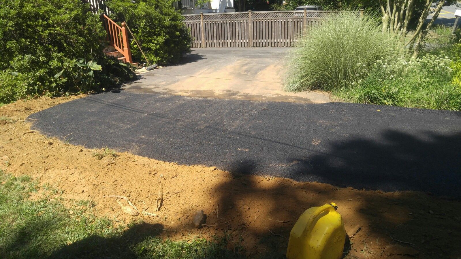 A fresh strip of black asphalt has been laid down on a dirt driveway bordered by a lawn and a wooden fence.