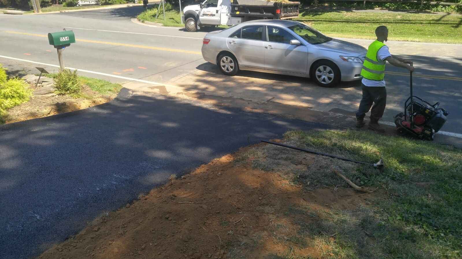 A worker in a high-visibility vest uses a plate compactor to smooth fresh asphalt on a driveway near a parked car.