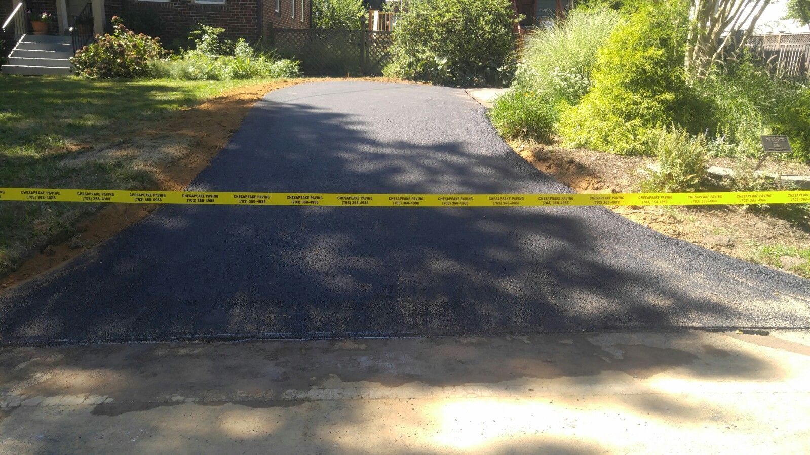 Freshly paved, dark gravel driveway bordered by grass and landscaping, blocked off by yellow caution tape.