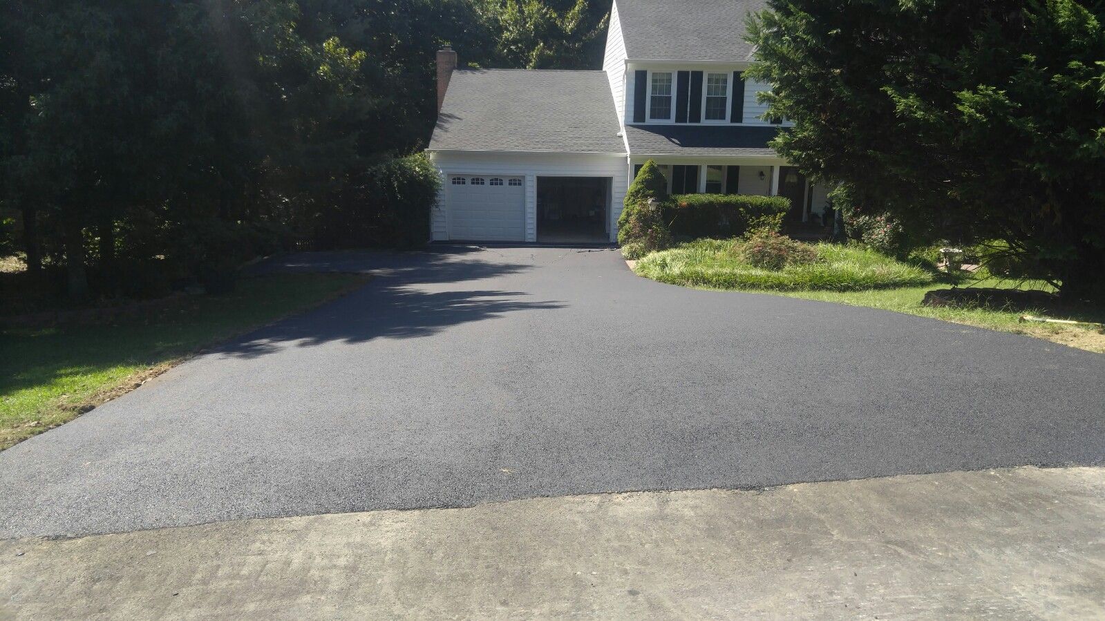 A freshly paved dark asphalt driveway leading up to a two-story house with a white garage and green trees.
