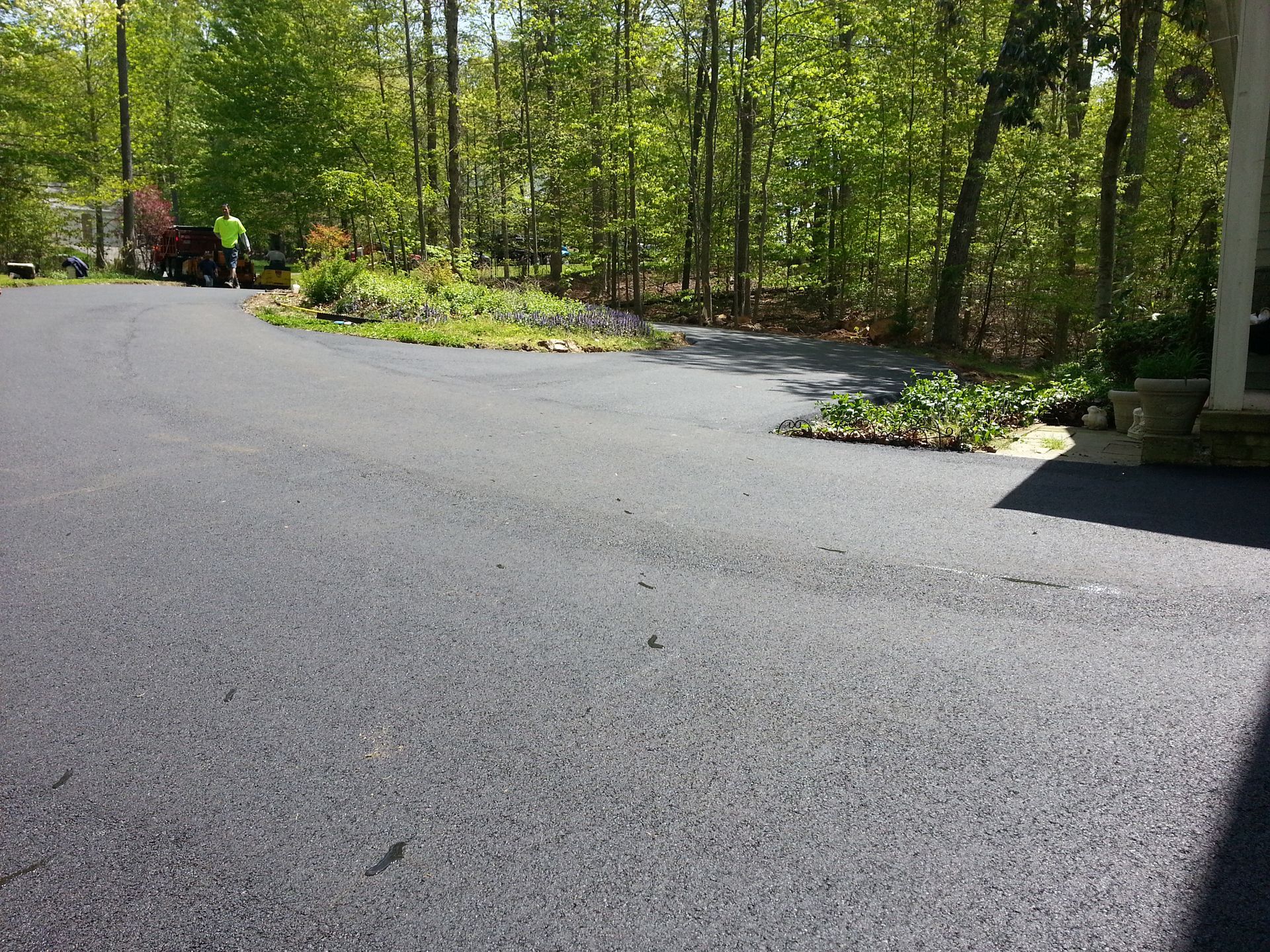 A fresh, dark asphalt driveway curves toward a wooded area with a small patch of green landscaping and a utility vehicle.