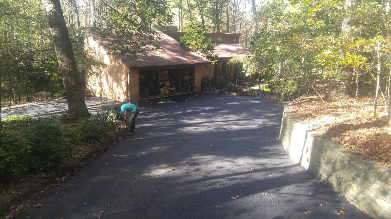 A person in a blue shirt works along the edge of a newly paved asphalt driveway in front of a house in a wooded area.