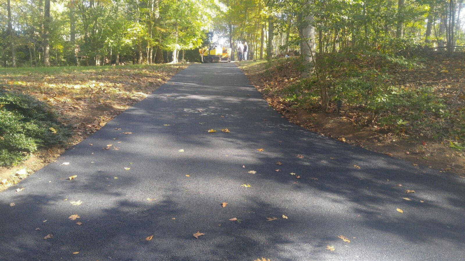 A newly paved dark asphalt path winds uphill through a wooded area with trees and scattered fallen leaves.