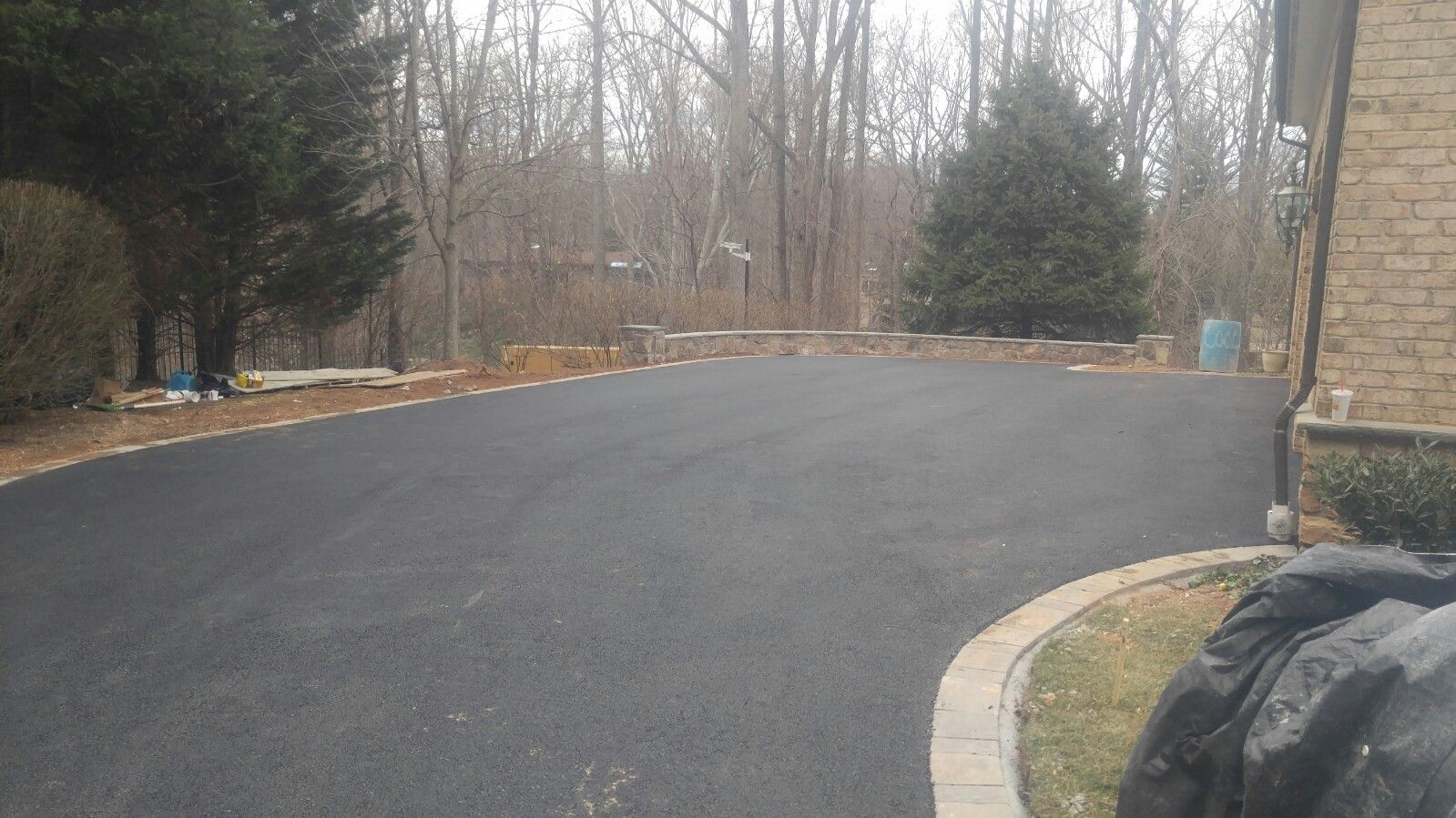 A freshly paved black asphalt driveway curves toward a brick home, bordered by a stone edge and a wooded background.