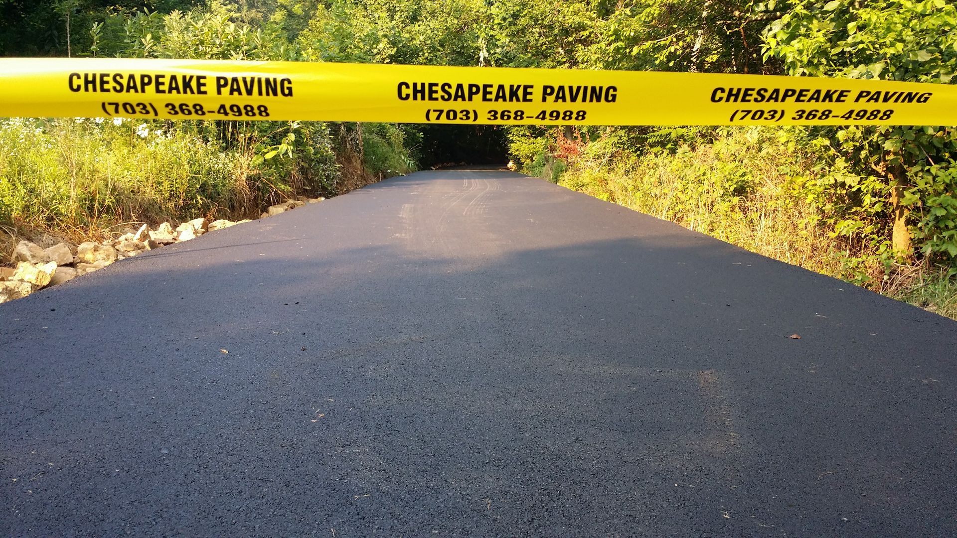 A freshly paved, dark asphalt path in a wooded area, blocked by yellow 