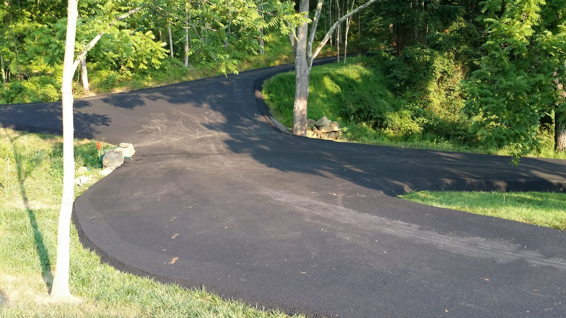 A freshly paved black asphalt driveway splits into two directions against a backdrop of green trees and grass.
