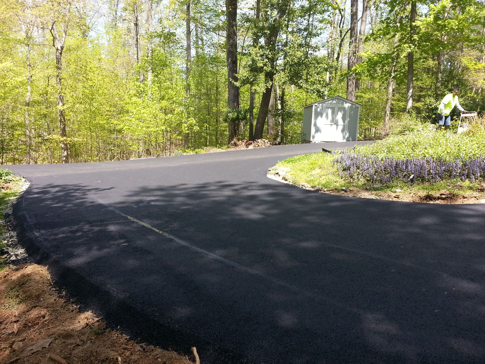 A newly paved black asphalt driveway curves through a sunlit wooded area with a small utility box in the background.