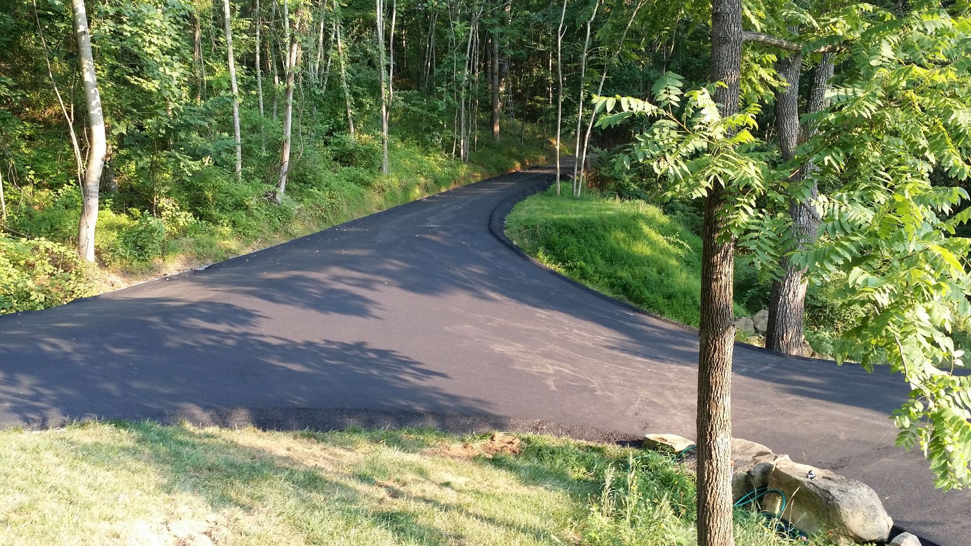 A newly paved asphalt driveway curves through a wooded area with surrounding green grass and trees.