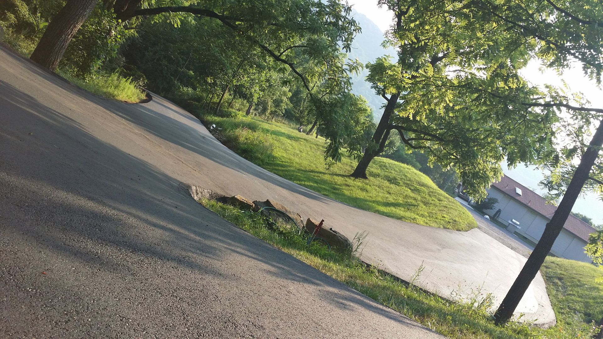 A paved road curves through a grassy, tree-lined landscape under bright sunlight, leading toward a building in the distance.