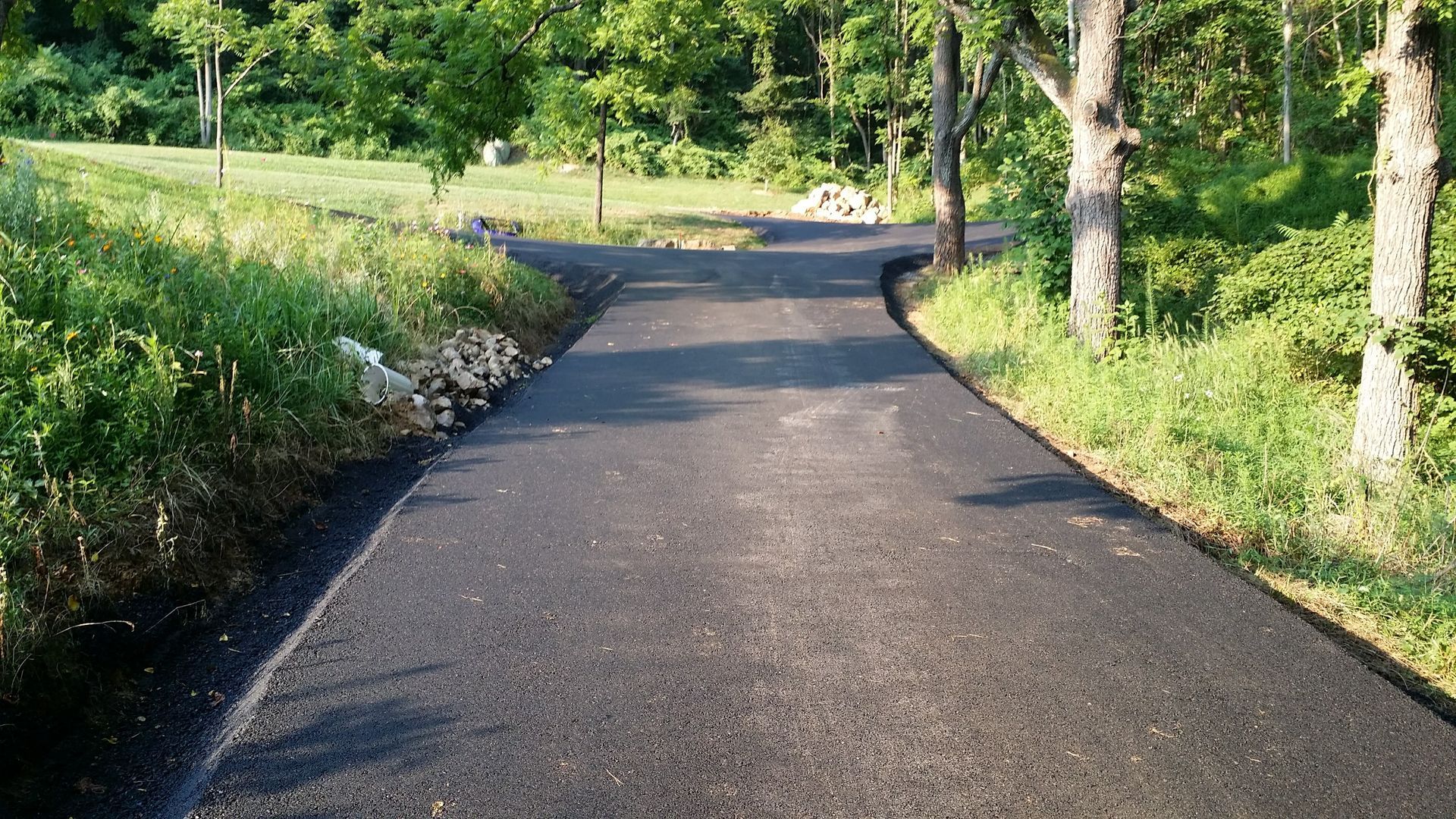 A newly paved asphalt path leads through a wooded area toward a Y-shaped junction surrounded by grass and trees.
