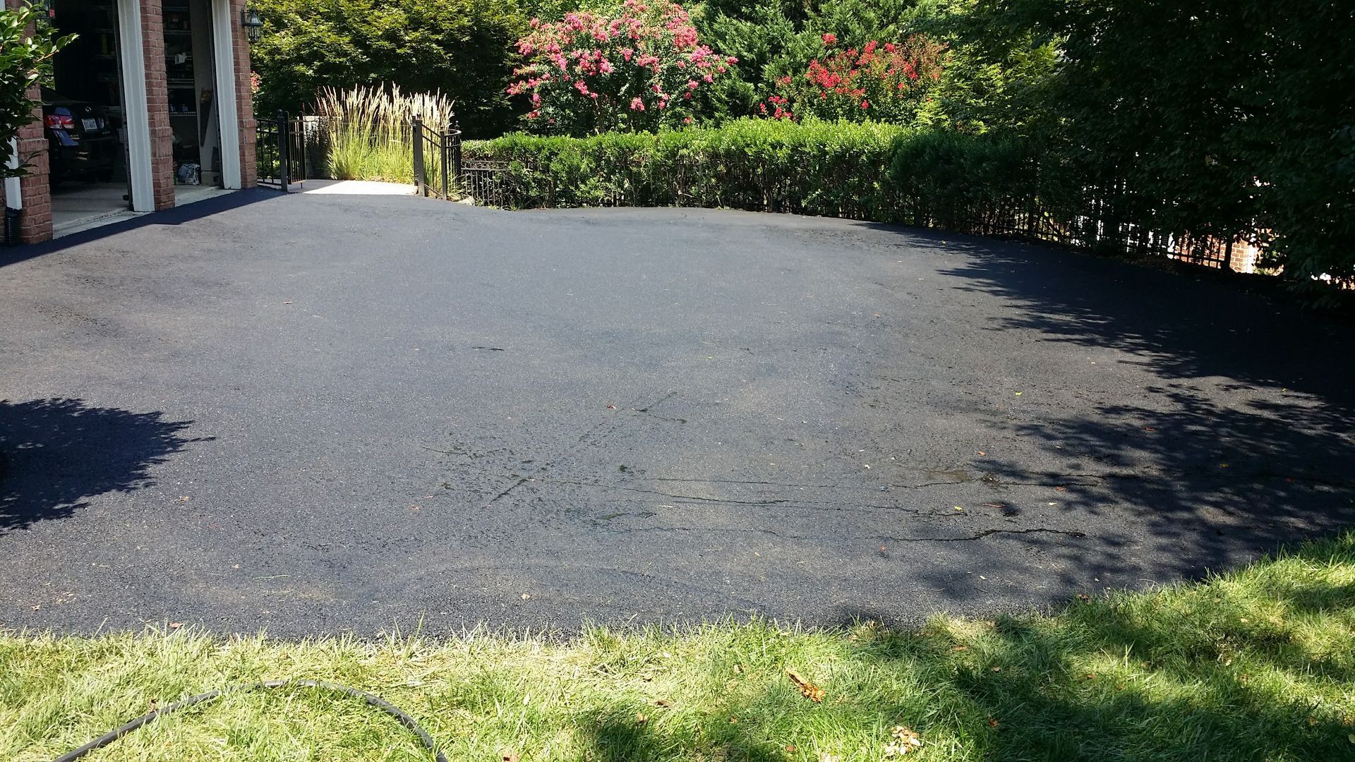 A freshly paved black asphalt driveway curves toward a garage, bordered by green grass and landscaping bushes.