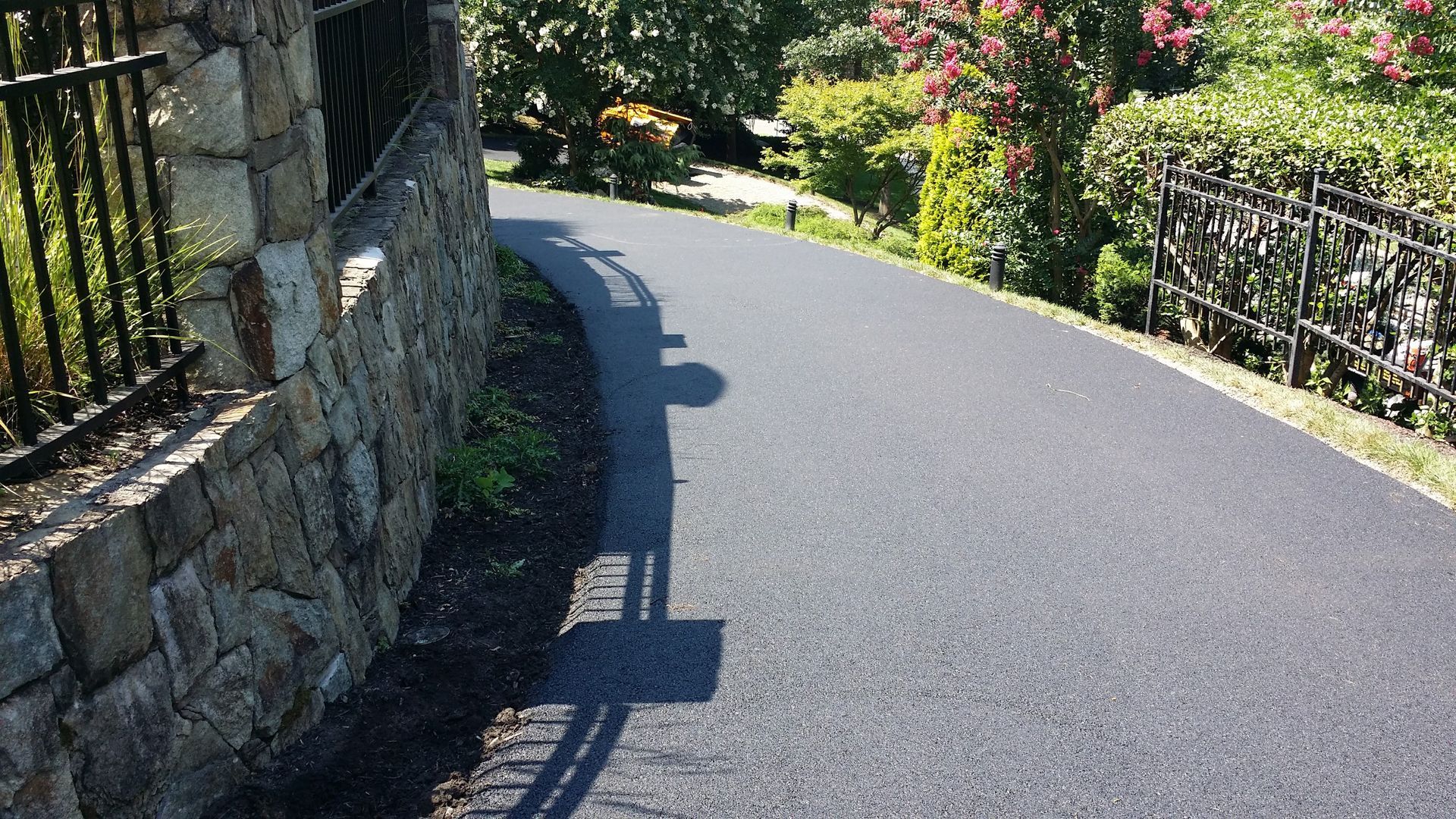 A smooth, dark asphalt driveway curves past a decorative stone retaining wall and lush green landscaping.