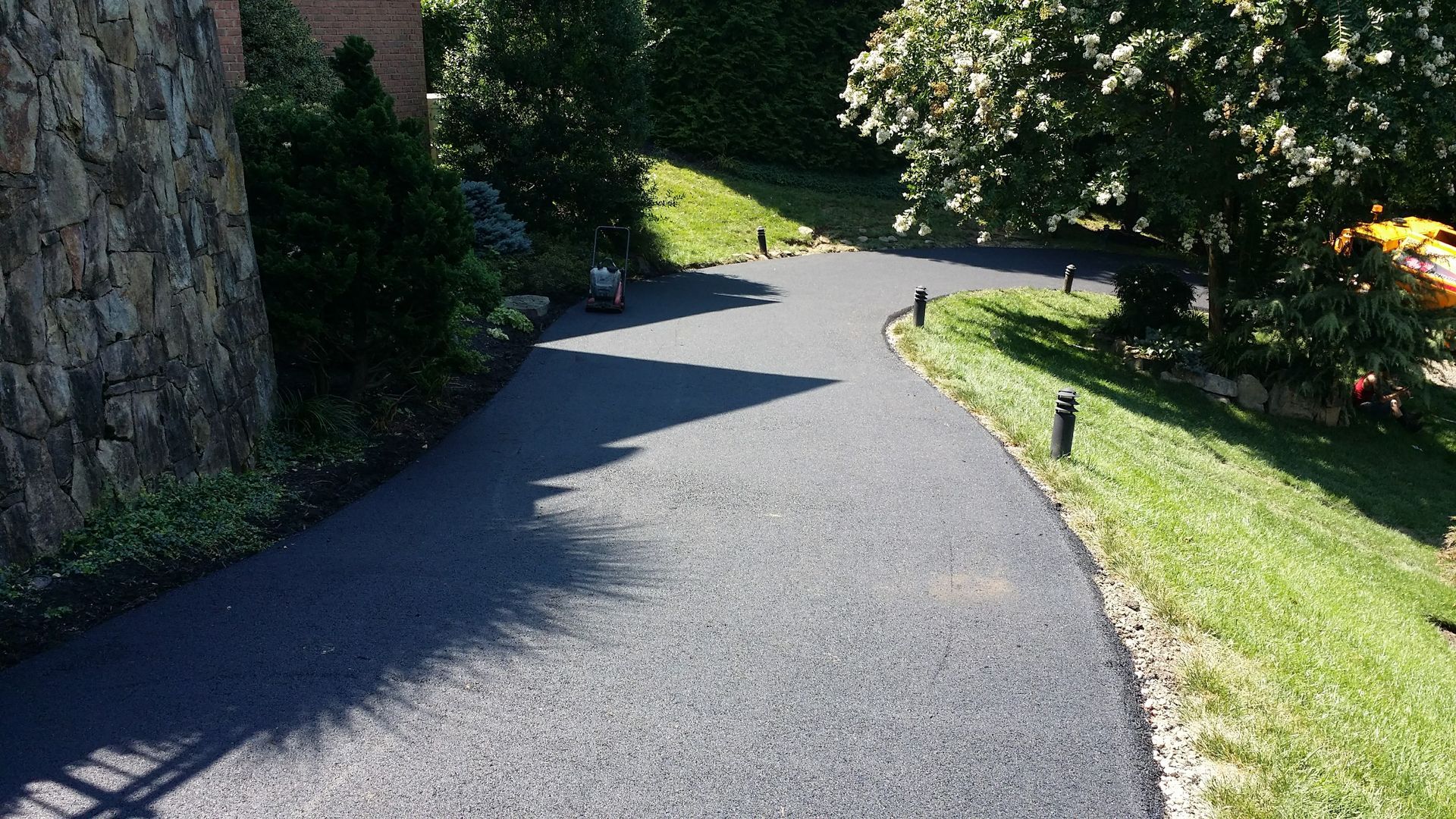 A freshly paved asphalt driveway curves past a stone wall and a grassy lawn with landscape lighting.