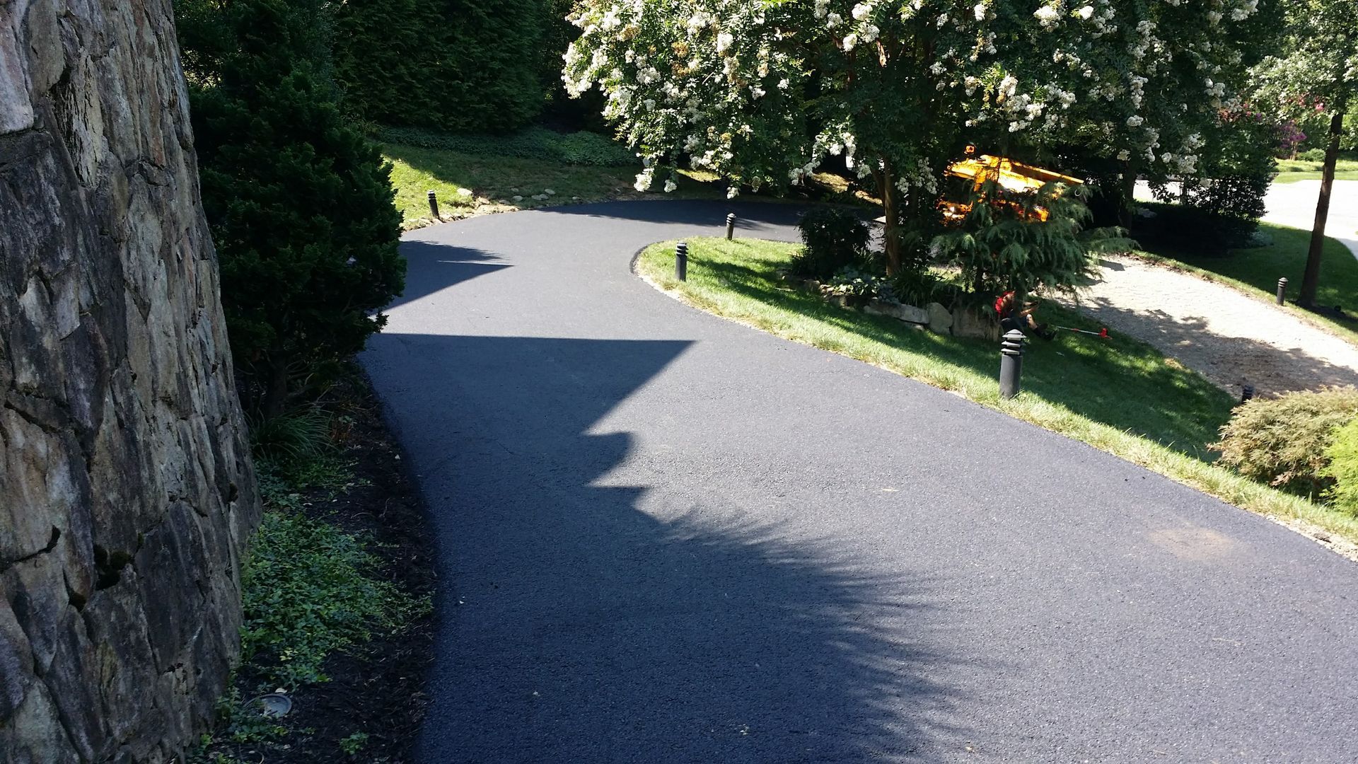 A freshly paved black asphalt driveway curves through a landscaped yard with green grass, trees, and stone accents.