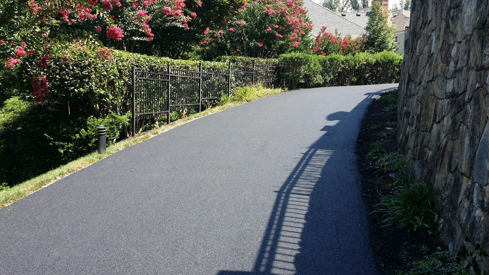 A paved driveway curves past a stone retaining wall and a hedge with blooming pink crape myrtle trees.