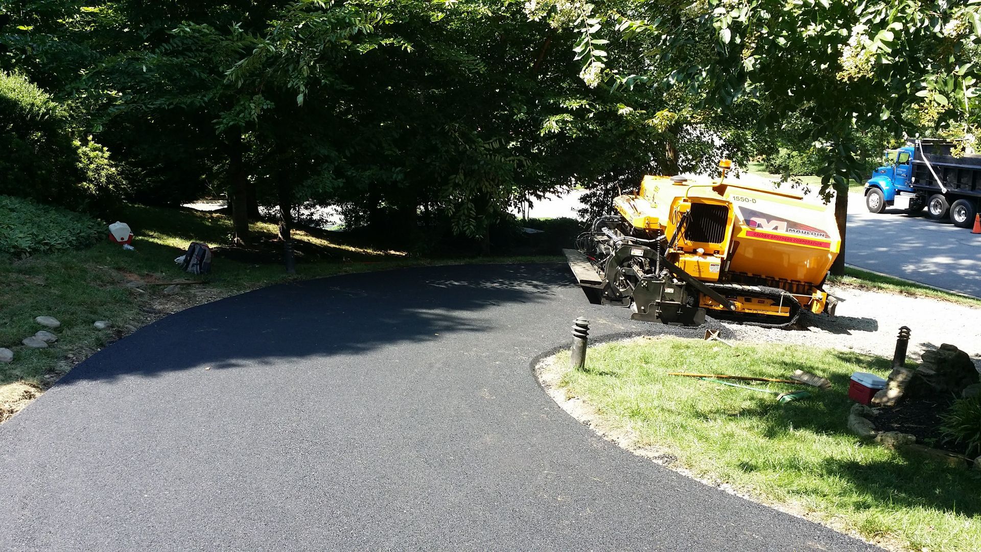 A yellow asphalt paver sits at the edge of a newly paved black driveway next to a grassy yard and trees.
