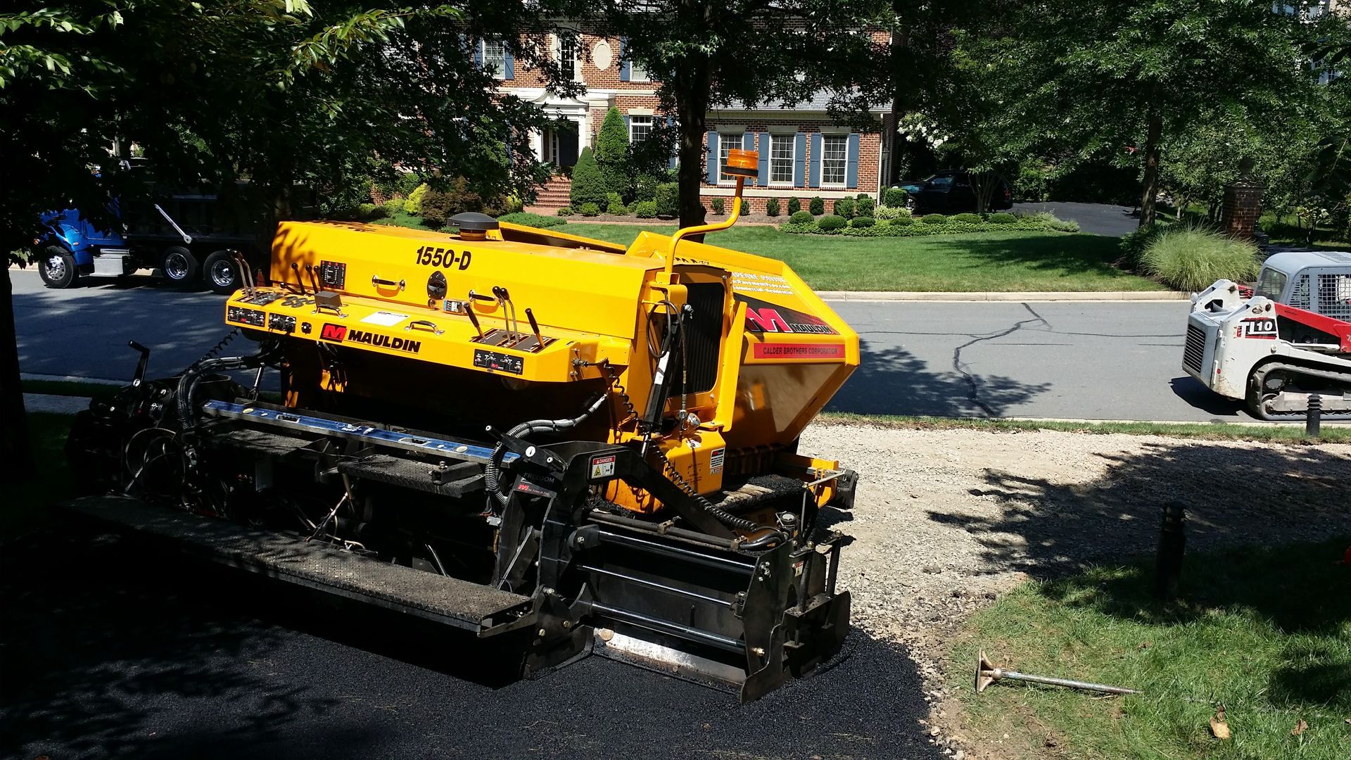 A bright yellow asphalt paver sits on a residential driveway near a suburban street.