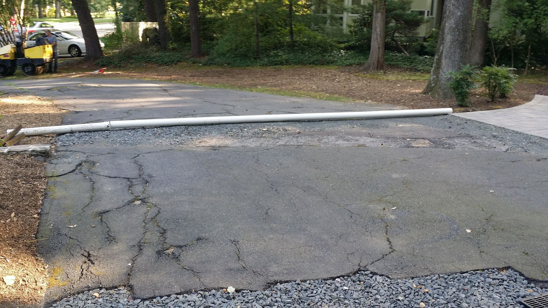 A white pipe lies across a cracked, weathered asphalt driveway near trees and a parked vehicle.