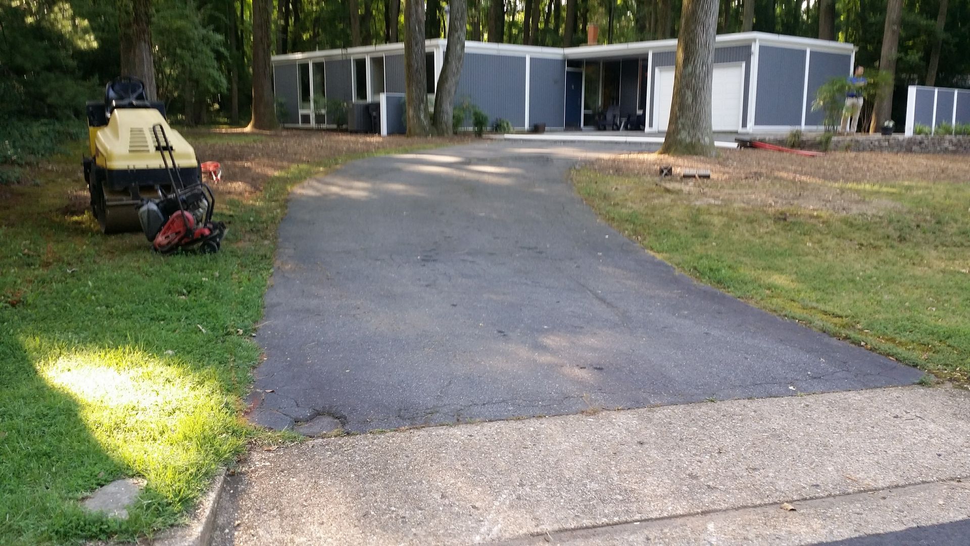 A driveway with fresh asphalt leads to a modern, gray single-story home nestled in a wooded yard with construction tools.
