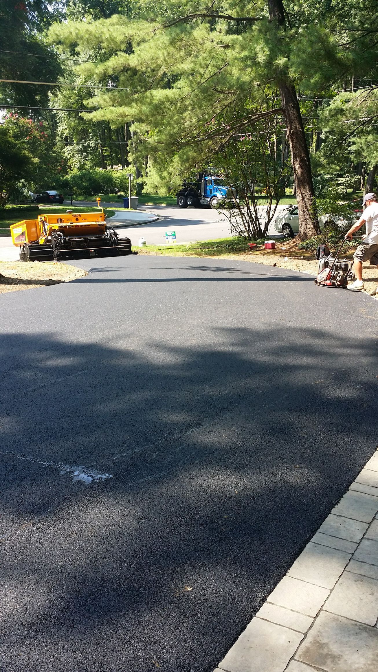 A person operates a machine to smooth newly laid asphalt on a residential driveway near a brick border.