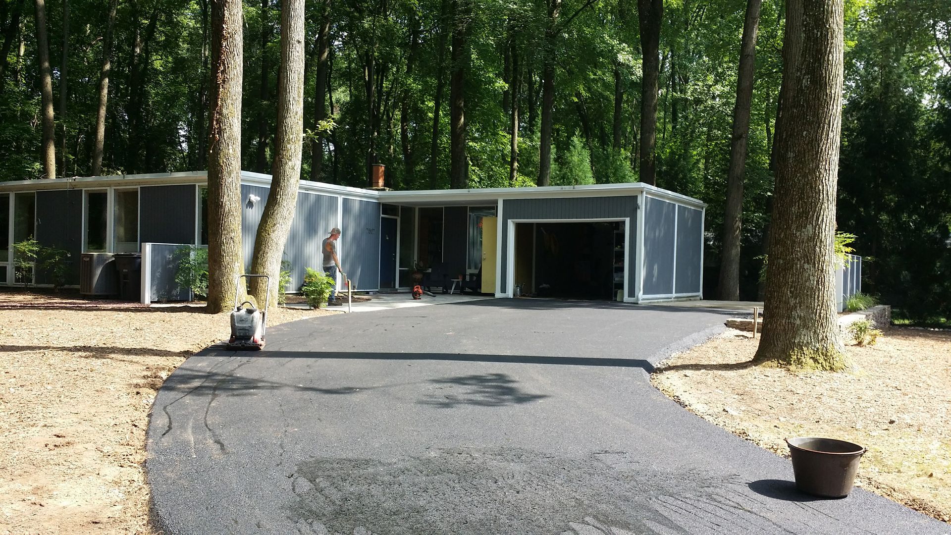 A gray, single-story house in a wooded area with a freshly paved asphalt driveway leading to a garage.