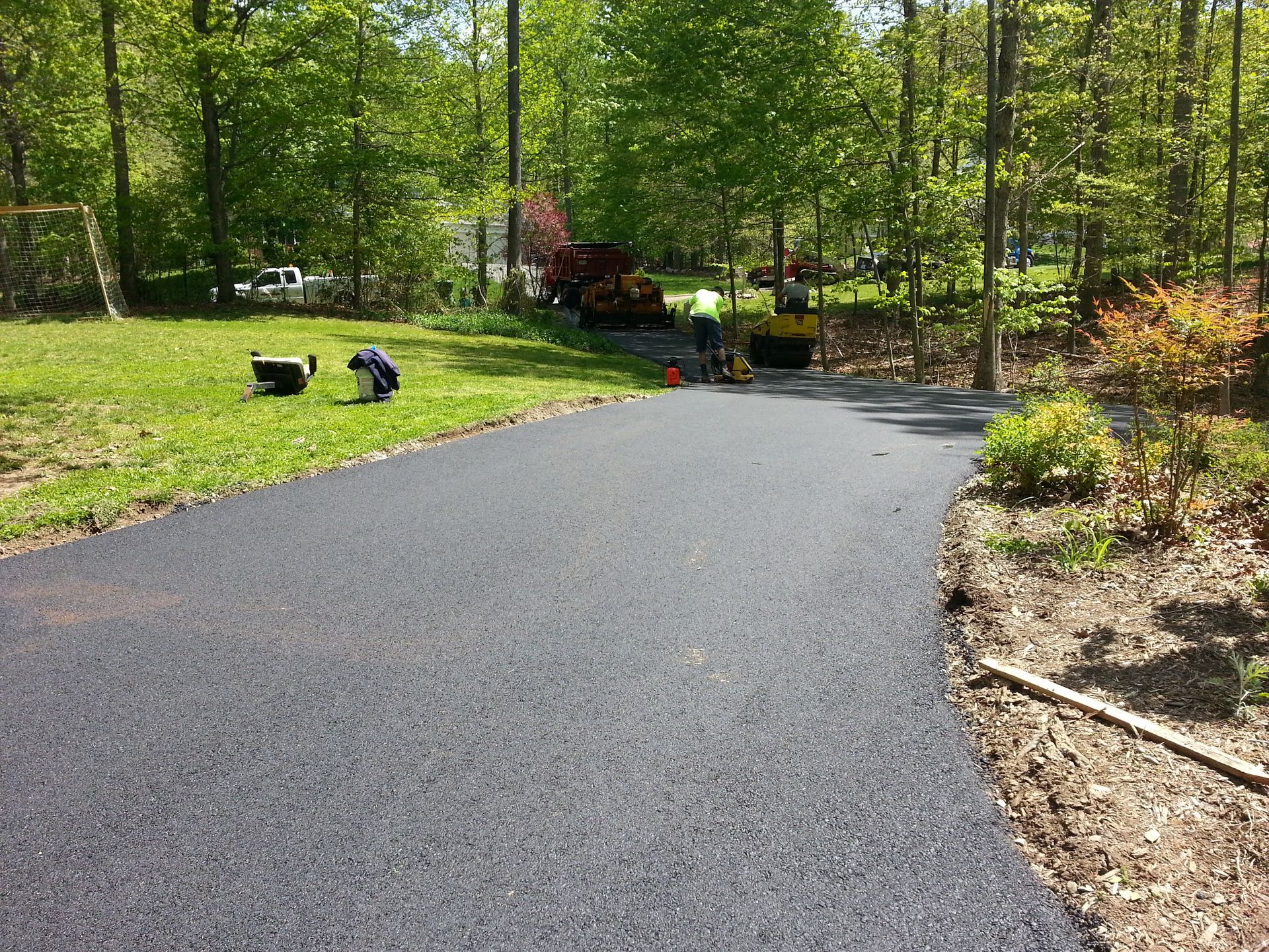 A freshly paved asphalt driveway extending into a wooded yard with a worker and machinery in the background.