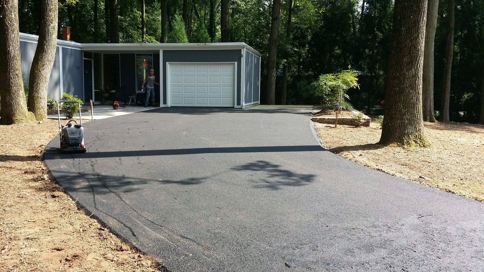 A newly paved asphalt driveway leads to a gray house with a white garage door, situated among tall trees in a wooded yard.