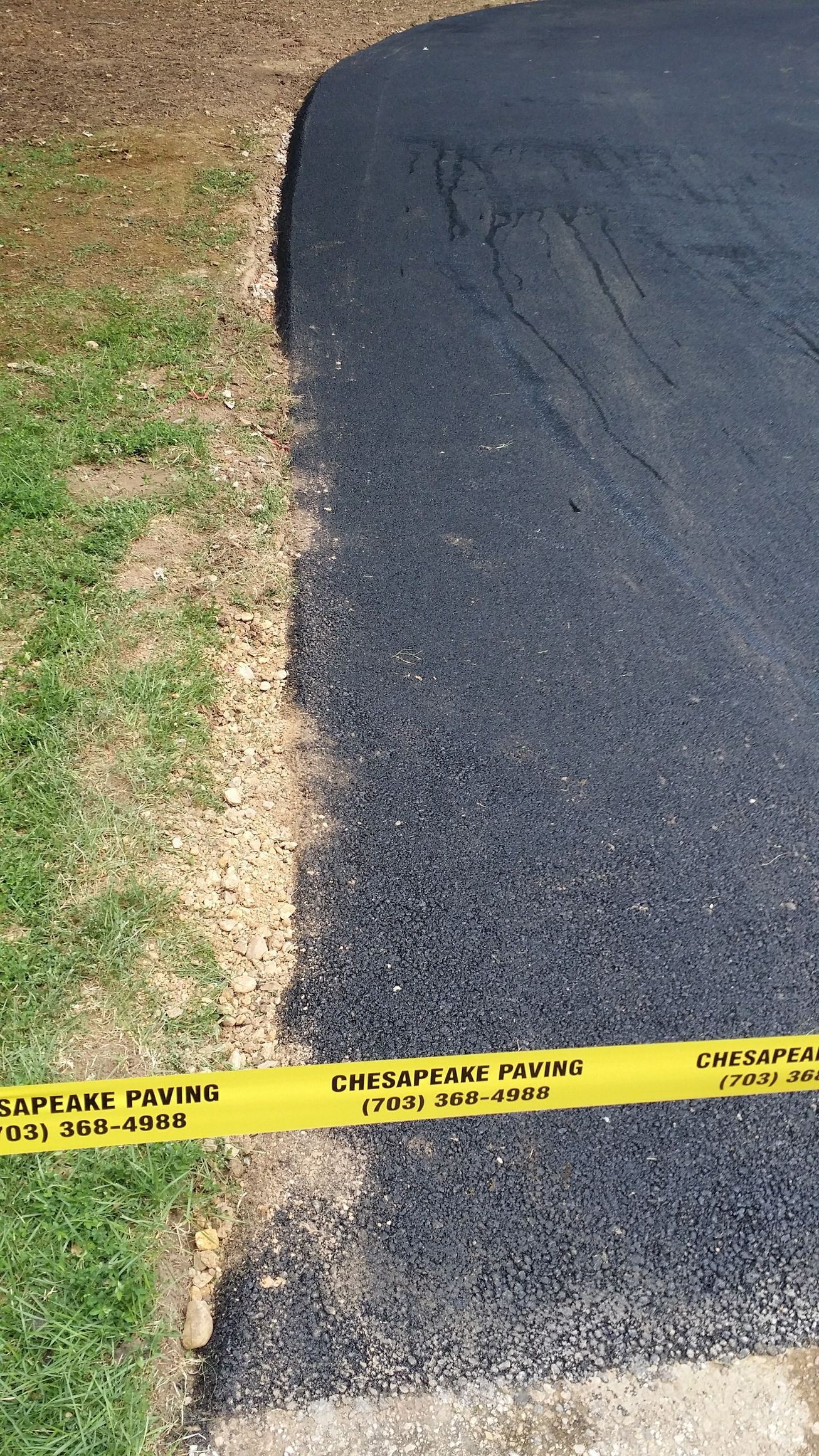 A patch of newly laid black asphalt driveway bordered by grass, with a yellow caution tape strung across the bottom.