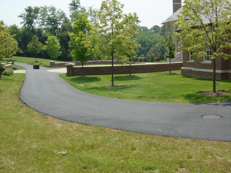 A wide, smooth asphalt driveway curves past a grassy lawn toward a brick building on a sunny day.