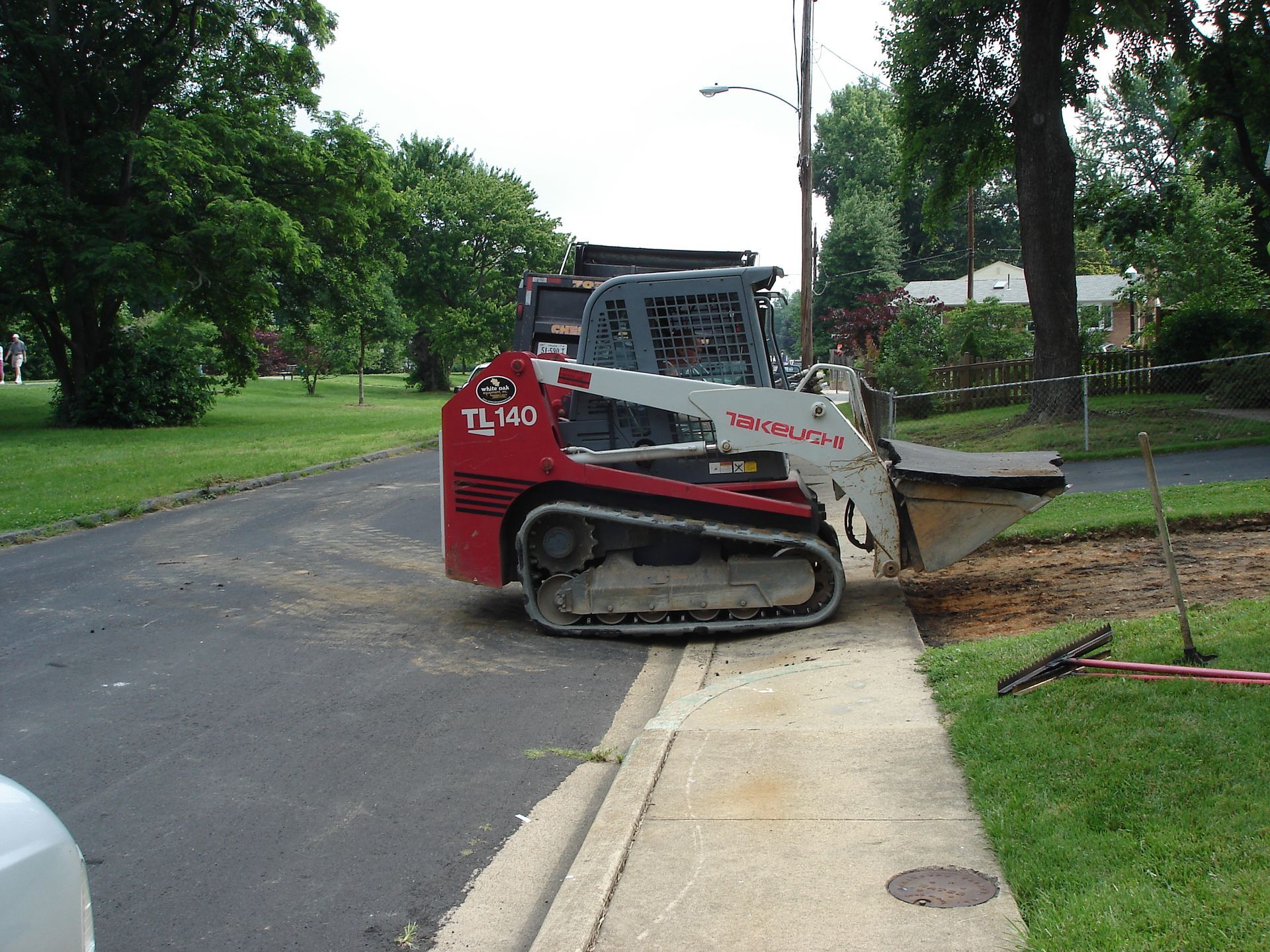 A red and white Takeuchi track loader sits at the edge of a paved road next to a sidewalk and grassy yard.