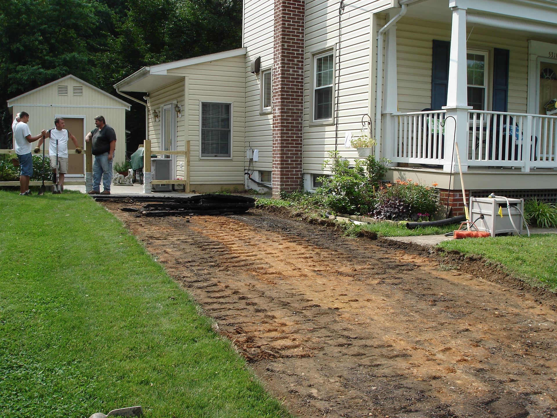 Three people stand on a residential property near a freshly excavated dirt driveway in front of a house.