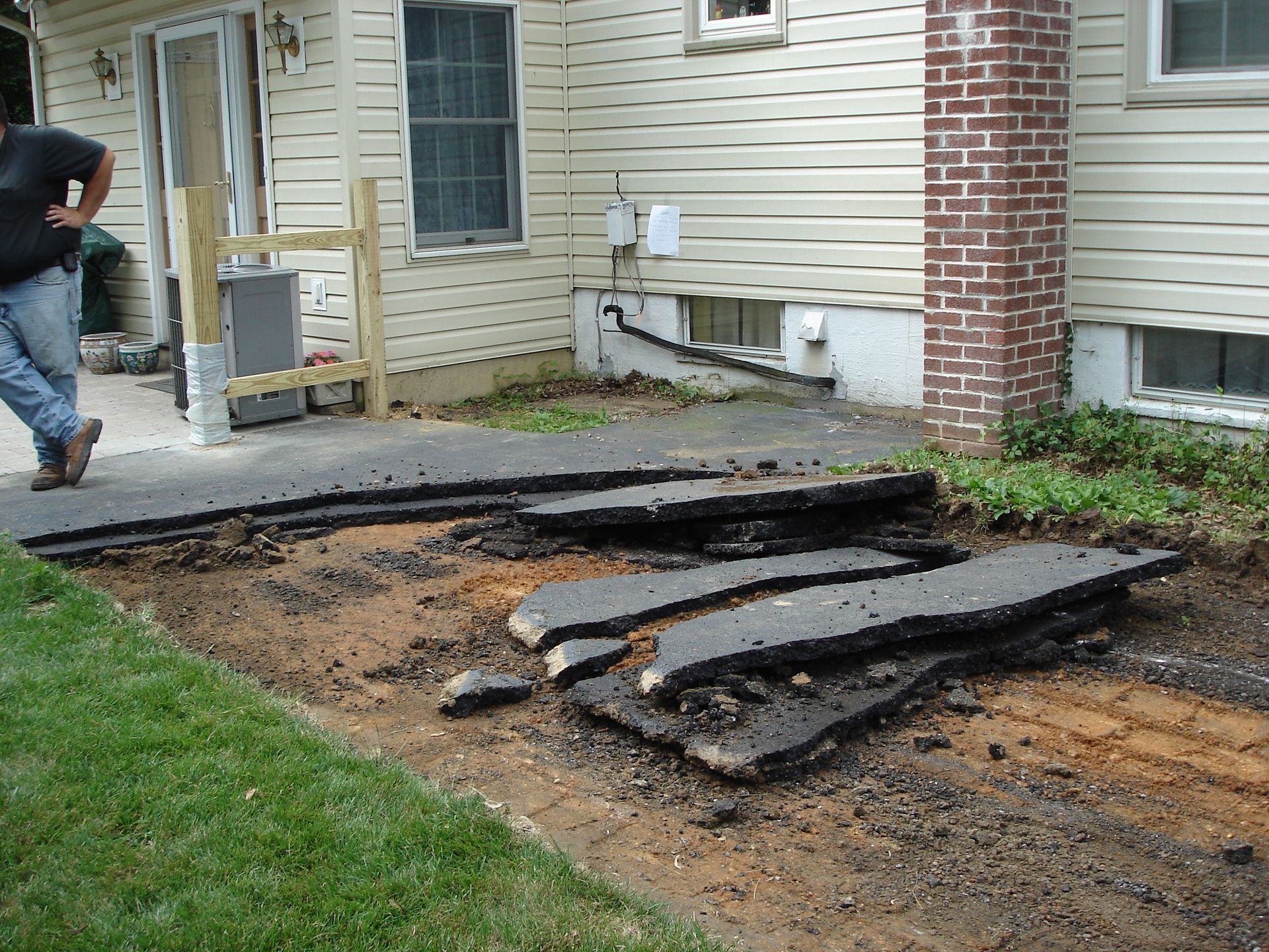 A worker stands beside a section of driveway with broken asphalt slabs removed to expose the soil underneath.