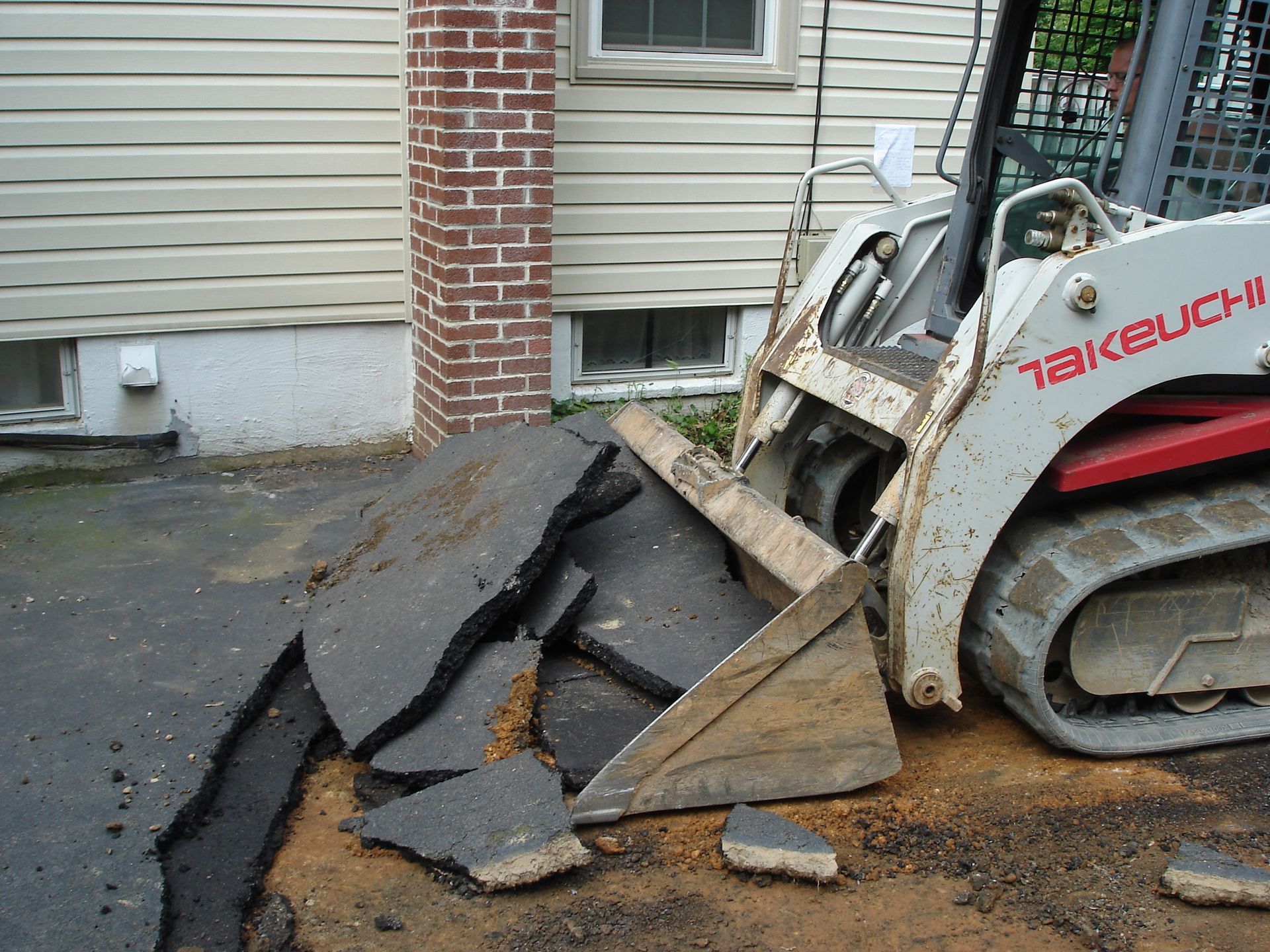 A white Takeuchi compact track loader breaking up an asphalt driveway next to a house with beige siding and a brick chimney.